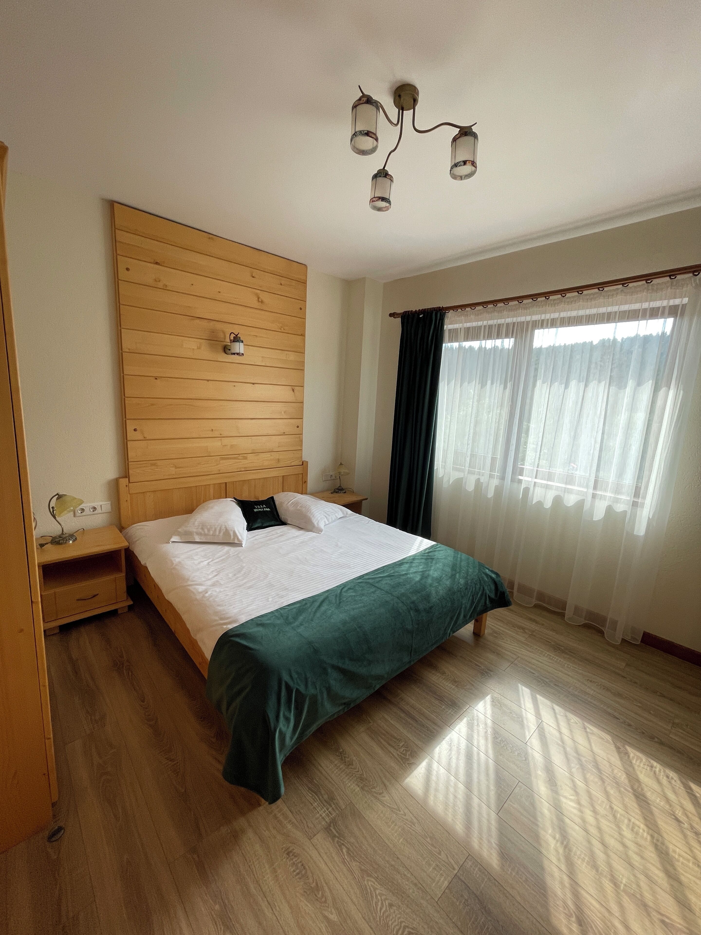A neatly arranged bedroom with a wooden headboard, a bed with white linens, a green throw blanket, a nightstand with a lamp, and a window with sheer curtains.