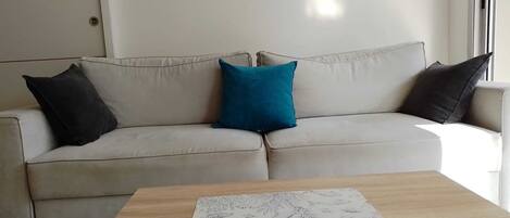 A beige sofa with two black and one teal pillow, a wooden coffee table with a printed coaster, and a window with blinds.