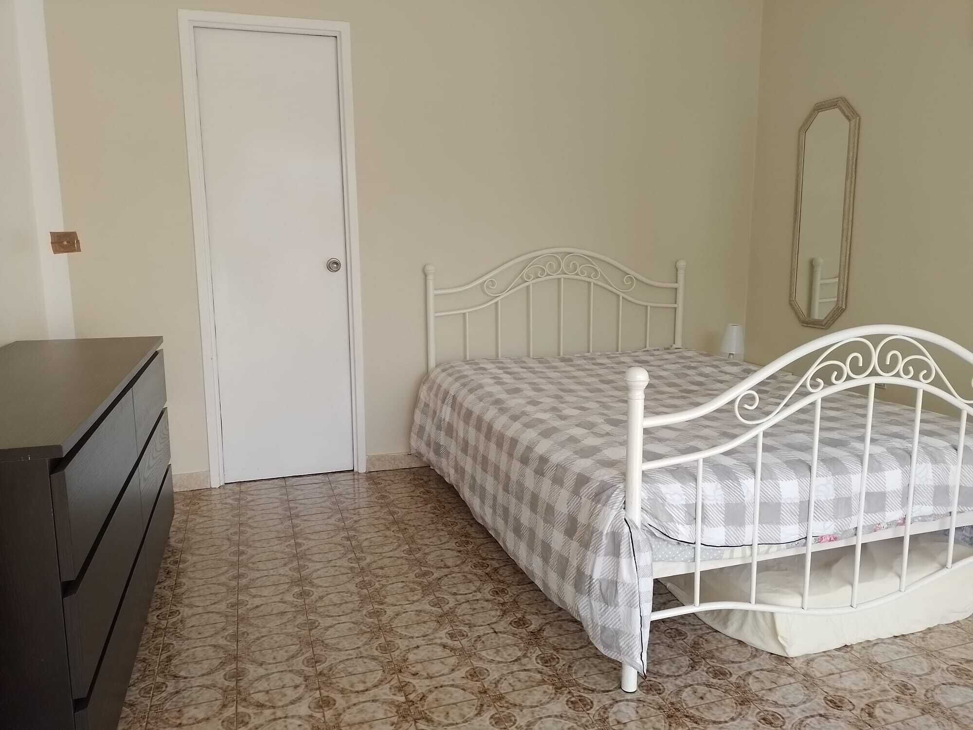 A bedroom with a white metal bed frame, a checkered bedspread, a dark wooden dresser, and patterned floor tiles.
