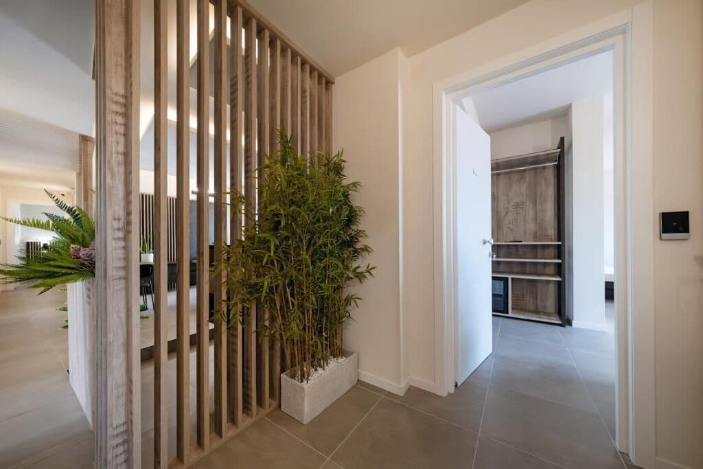 Un pasillo con un separador de listones de madera, una planta de bambú en maceta y vista a un cuarto con puerta de madera y estanterías.