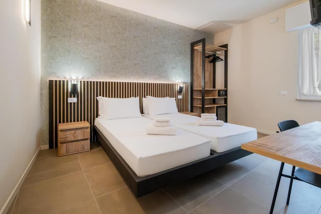 A modern hotel room with a large bed, a wooden nightstand, a built-in wardrobe, and a dining table.