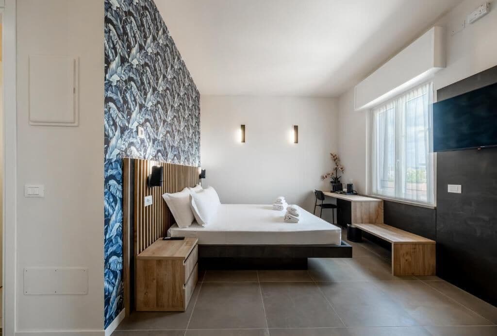 A modern hotel room with a bed, bedside tables, a bench, a desk, and a large window.
