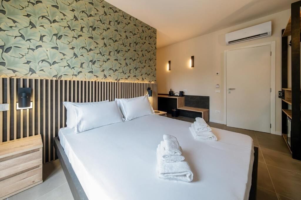 A modern hotel room with a large bed, a wooden headboard, a wall with a leaf pattern, a bedside table, a nightstand, a TV, and an air conditioning unit.