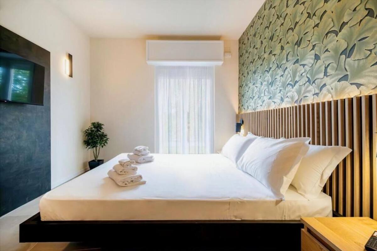 Una habitación de hotel moderna con una cama, un televisor fijado a la pared, una planta en maceta y una ventana con persianas.
