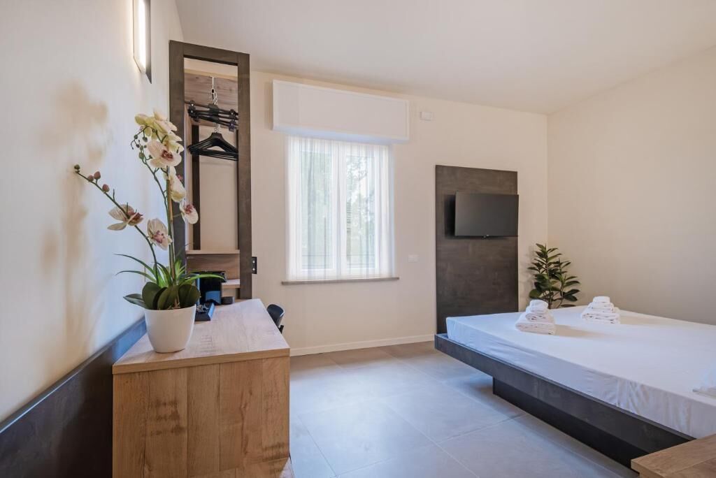 Un dormitorio moderno con cama, cómoda de madera, televisor montado en la pared y ventana con cortinas traslúcidas.