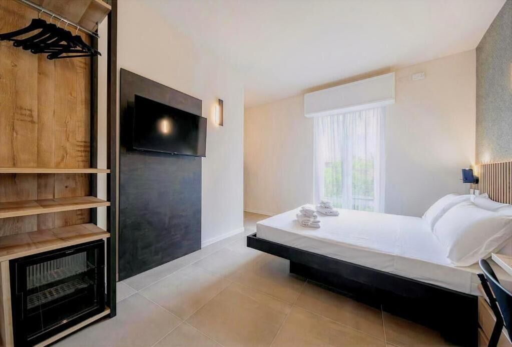 A modern hotel room with a bed, a flat-screen TV mounted on the wall, a wooden shelf unit, and a window with curtains.