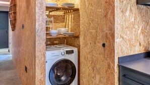 A compact kitchen with wooden walls, a built-in washing machine, and a shelf with dishes.