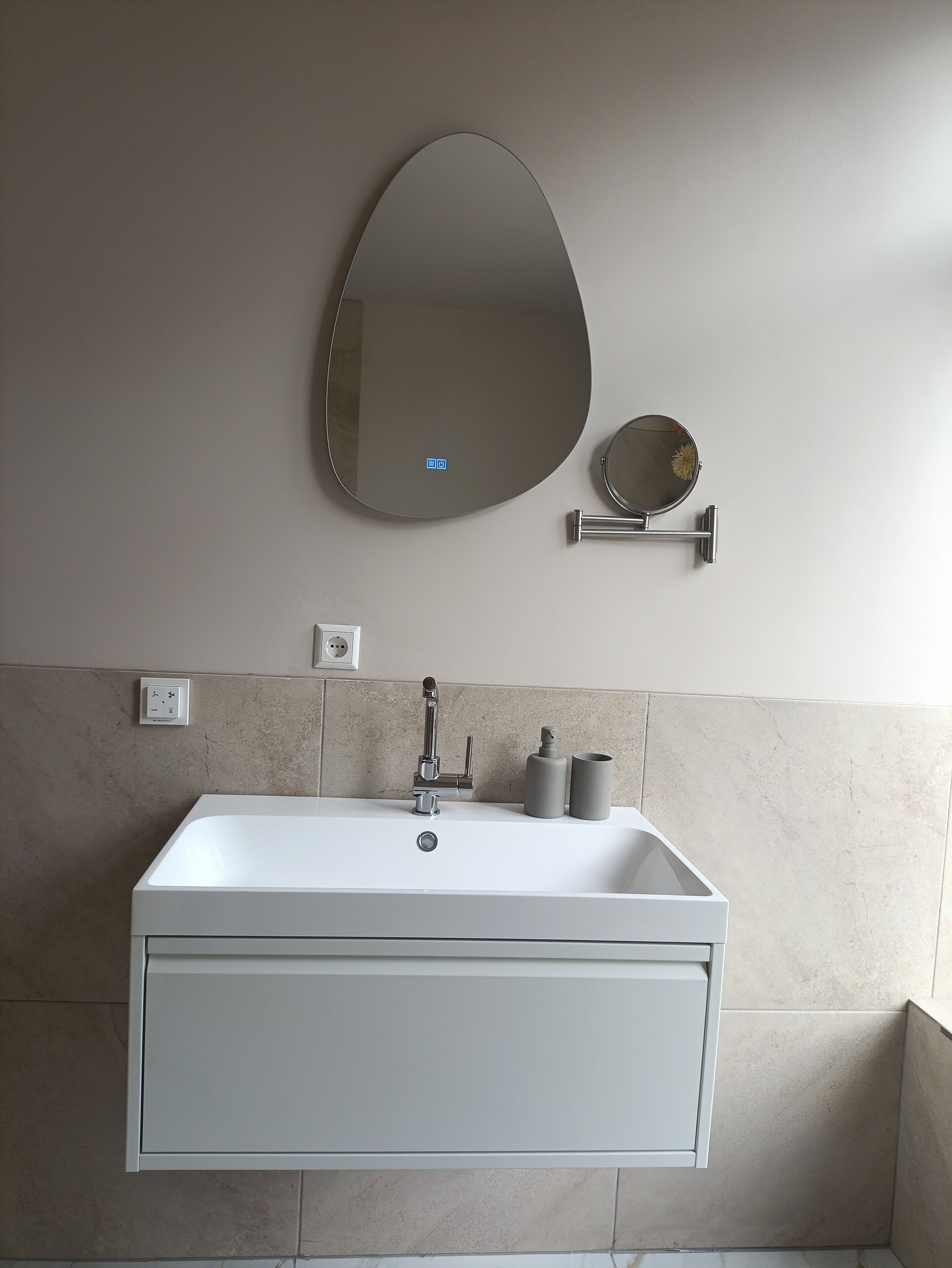 A bathroom with a white sink, a large oval mirror, a round mirror, and a towel dispenser.