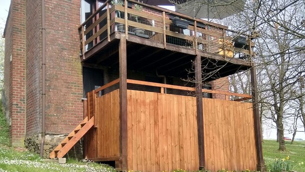 A rustic wooden structure with a balcony, a brick chimney, and a grassy area with flowers.