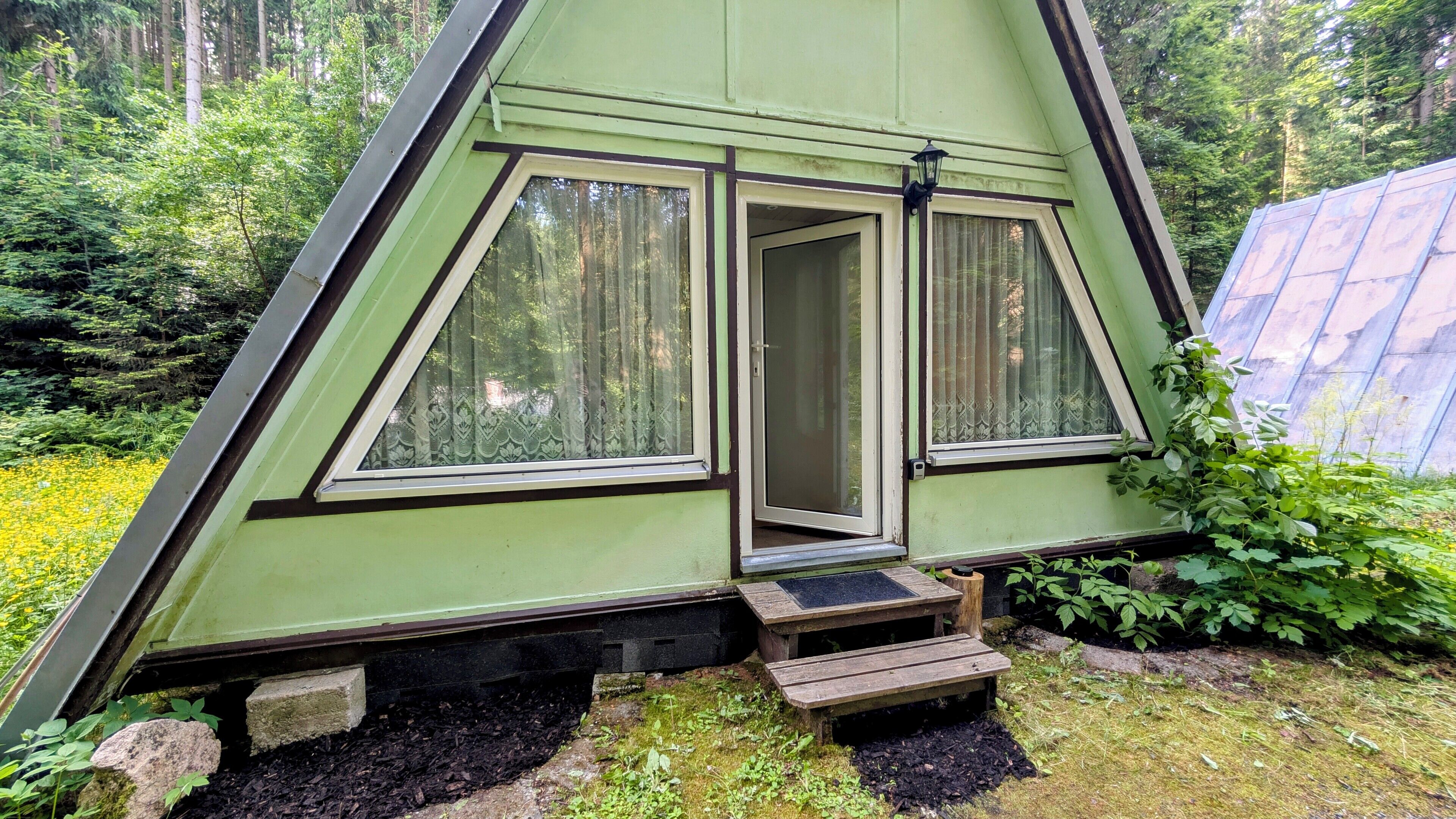 A triangular A-frame cabin with two large windows, a door, and a small wooden staircase leading to the entrance.