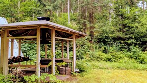 A wooden gazebo with benches and a table, surrounded by dense forest.