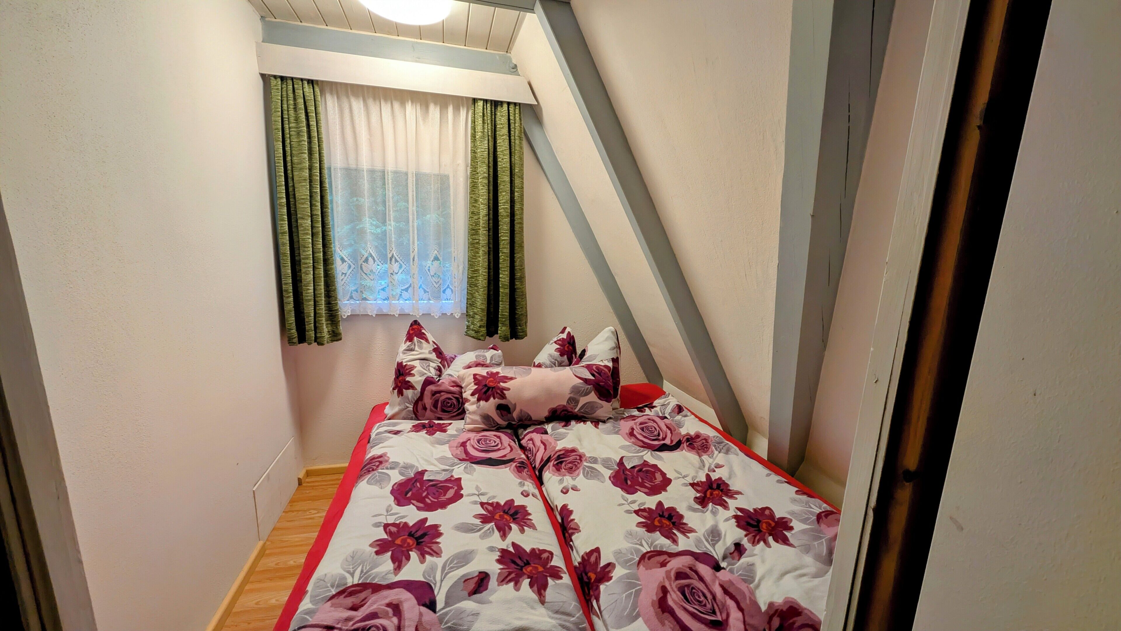 Ein kleiner, schmaler Raum mit einem Bett, einem Fenster mit Vorhängen und einer schrägen Decke.