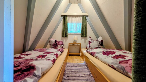 Una habitación pequeña y triangular con dos camas, una mesita de noche, una lámpara y una ventana con cortinas.
