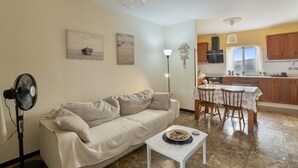A living room with a sofa, a fan, a table with a decorative bowl, a dining area with chairs and a table, and a kitchen with wooden cabinets and a window.