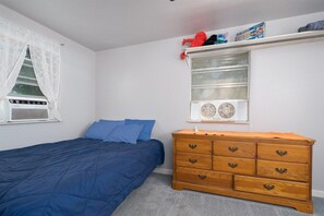 Un dormitorio con una cama, un aparador, una ventana con ventilador y un estante con juguetes.