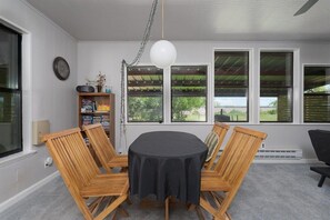 A dining area with a round table, four wooden chairs, a black tablecloth, a clock on the wall, and a window with a view of greenery.