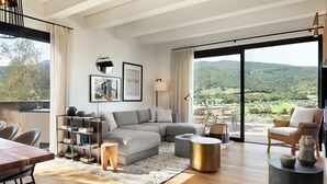 A modern living room with a sectional sofa, a dining table, and a view of a lush landscape through large windows. - Argentario Golf Villas (Monte Argentario)