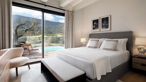 A bedroom with a large bed, a bench, a chair, and a view of a mountainous landscape through a sliding glass door. - Argentario Golf Villas (Monte Argentario)