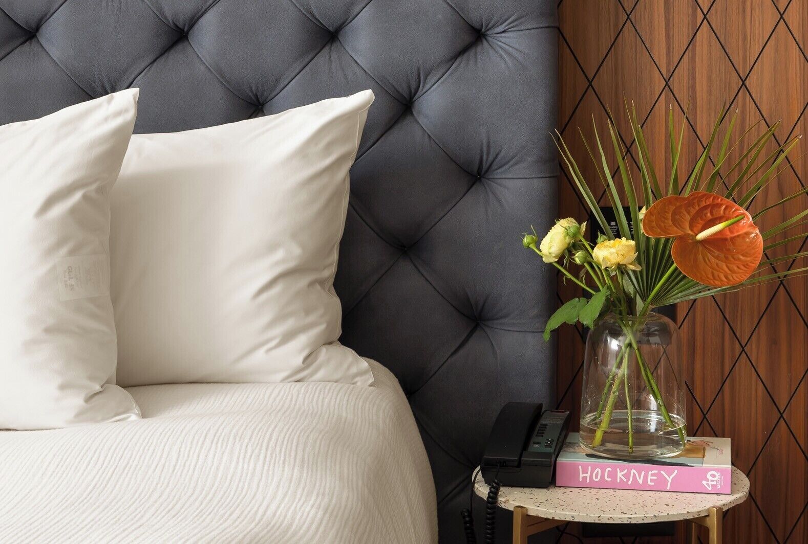 Un dormitorio con cabecera capitonada, ropa de cama blanca, una mesita de noche con un libro y un florero.