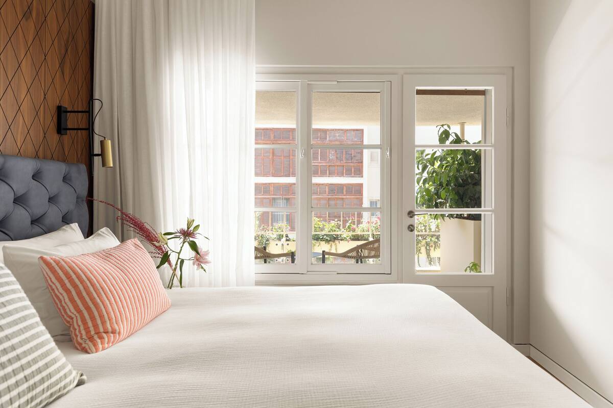A bedroom with a bed, a window with a view of buildings, and a plant on the windowsill.