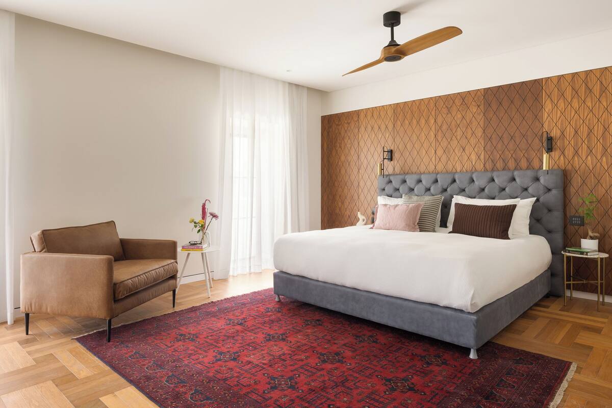 A modern bedroom with a large bed, a sofa, a red patterned rug, and a wooden accent wall.