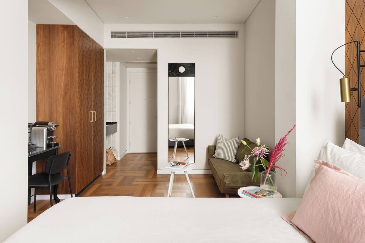 A modern hotel room with a wooden wardrobe, a dining area with a table and chairs, a mirror, a sofa with cushions, and a bedside table with a vase of flowers.