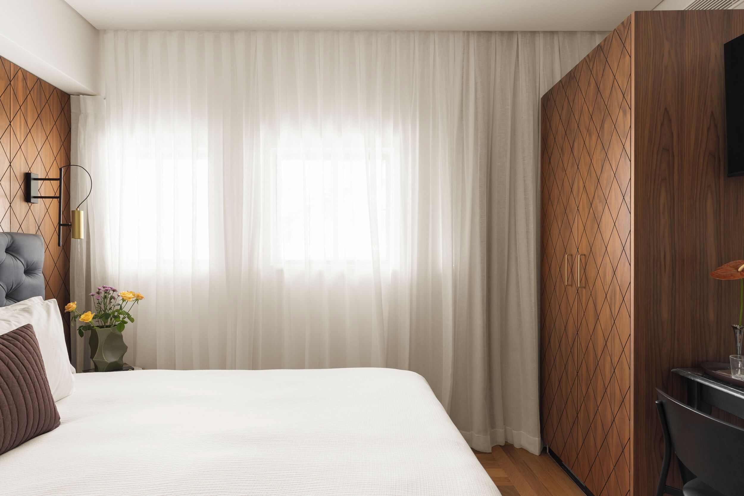 A hotel room with a large bed, a desk, a chair, a TV, and a window with sheer curtains.