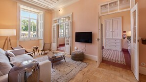 A living room with a sofa, coffee table, and chairs, a television mounted on the wall, and a large window with a view of the outdoors. - Charm & history in the heart of VRSA (Vila Real de Santo António)
