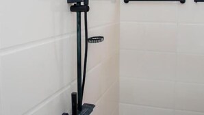 Une salle de bain avec des murs carrelés de carreaux blancs, une fenêtre, une douche avec un pommeau de douche amovible et des toilettes.