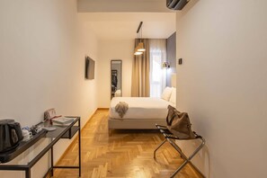 Quarto de hotel pequeno e funcional, com cama, escrivaninha, cadeira, espelho e janela com cortinas.