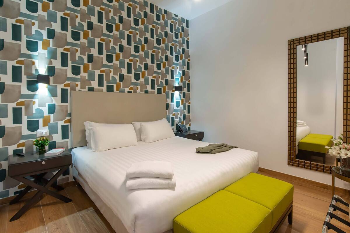 Una cama bien tendida con sábanas blancas y un banco verde, una mesita de noche con lámpara y un papel tapiz estampado.