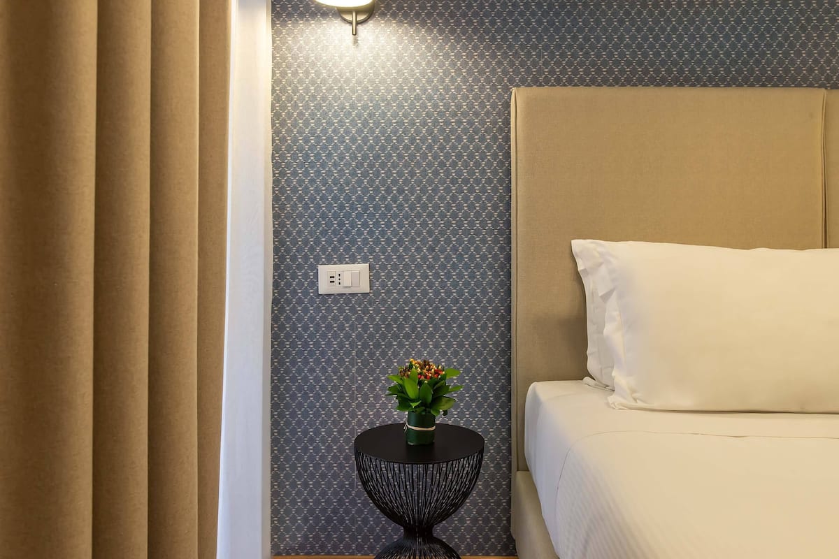 Habitación de hotel con una cama, una mesita de noche con un jarrón de flores y un papel tapiz estampado.