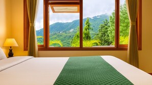 A hotel room with a bed, a lamp, and a window with a view of green hills and trees. - Hotel Shiva Lake - Near Mussoorie Lake (Mussoorie)