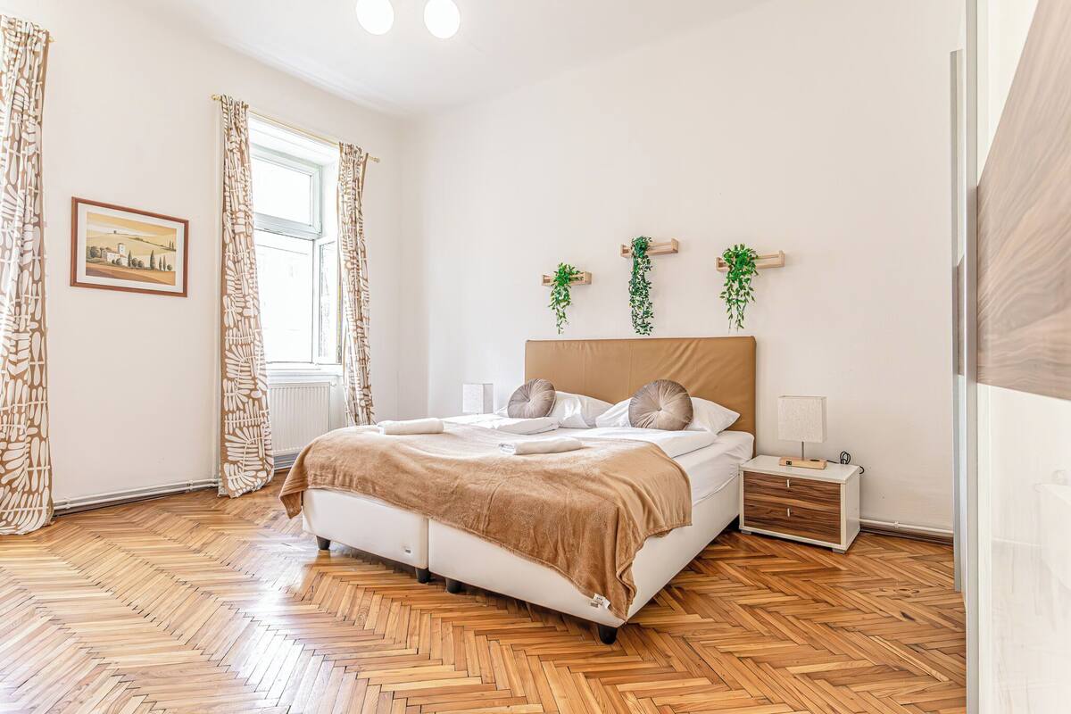 Un dormitorio con suelo de madera en patrón de espiga, una cama con manta marrón, una mesita de noche con lámpara y dos macetas colgantes en la pared.