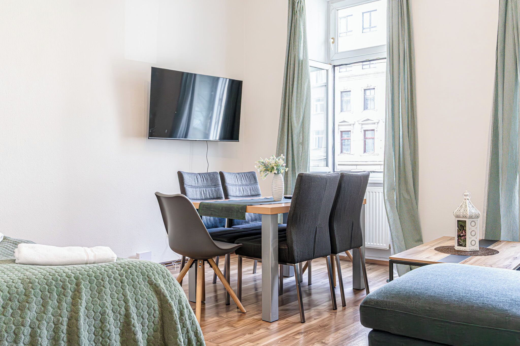 Une chambre d’hôtel moderne équipée d’un téléviseur à écran plat fixé au mur, d’une table à manger avec des chaises, d’un lit recouvert d’une couverture verte et d’une fenêtre avec des rideaux.
