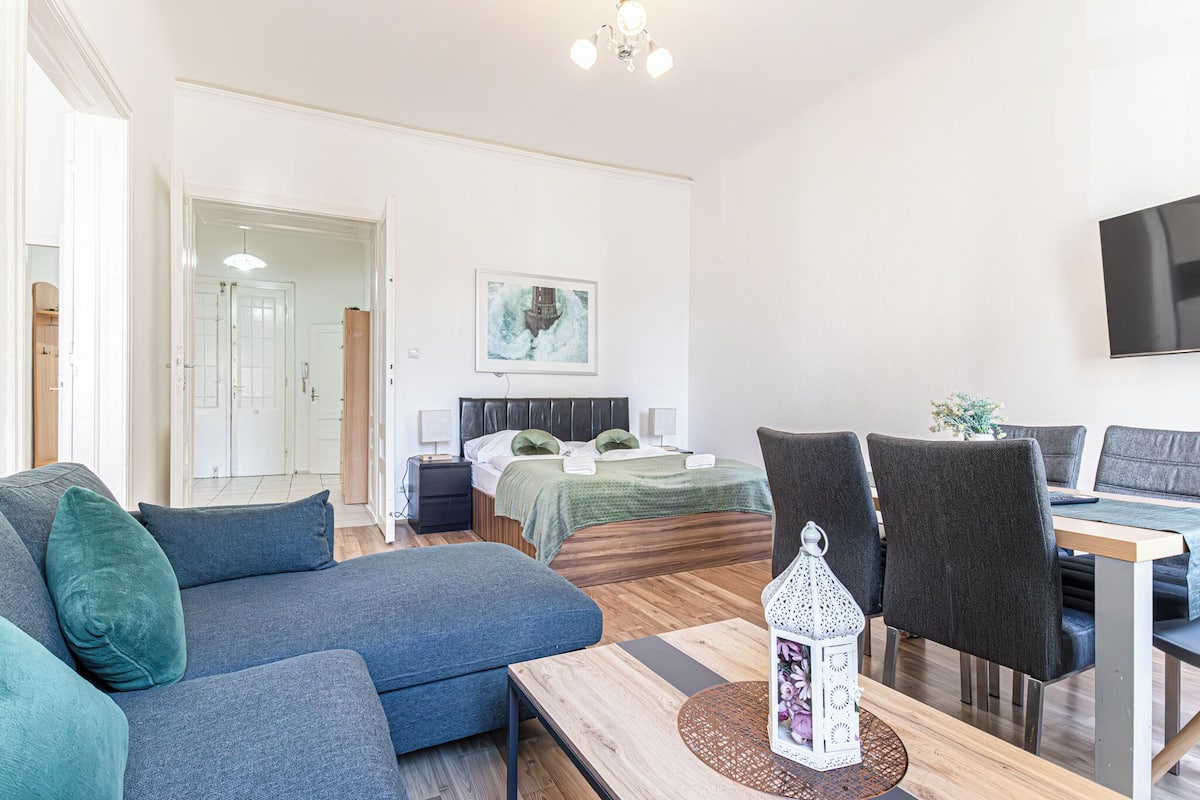 Una sala moderna con un sofá, una mesa de comedor y sillas. Hay una cama con colcha verde y un cuadro en la pared.