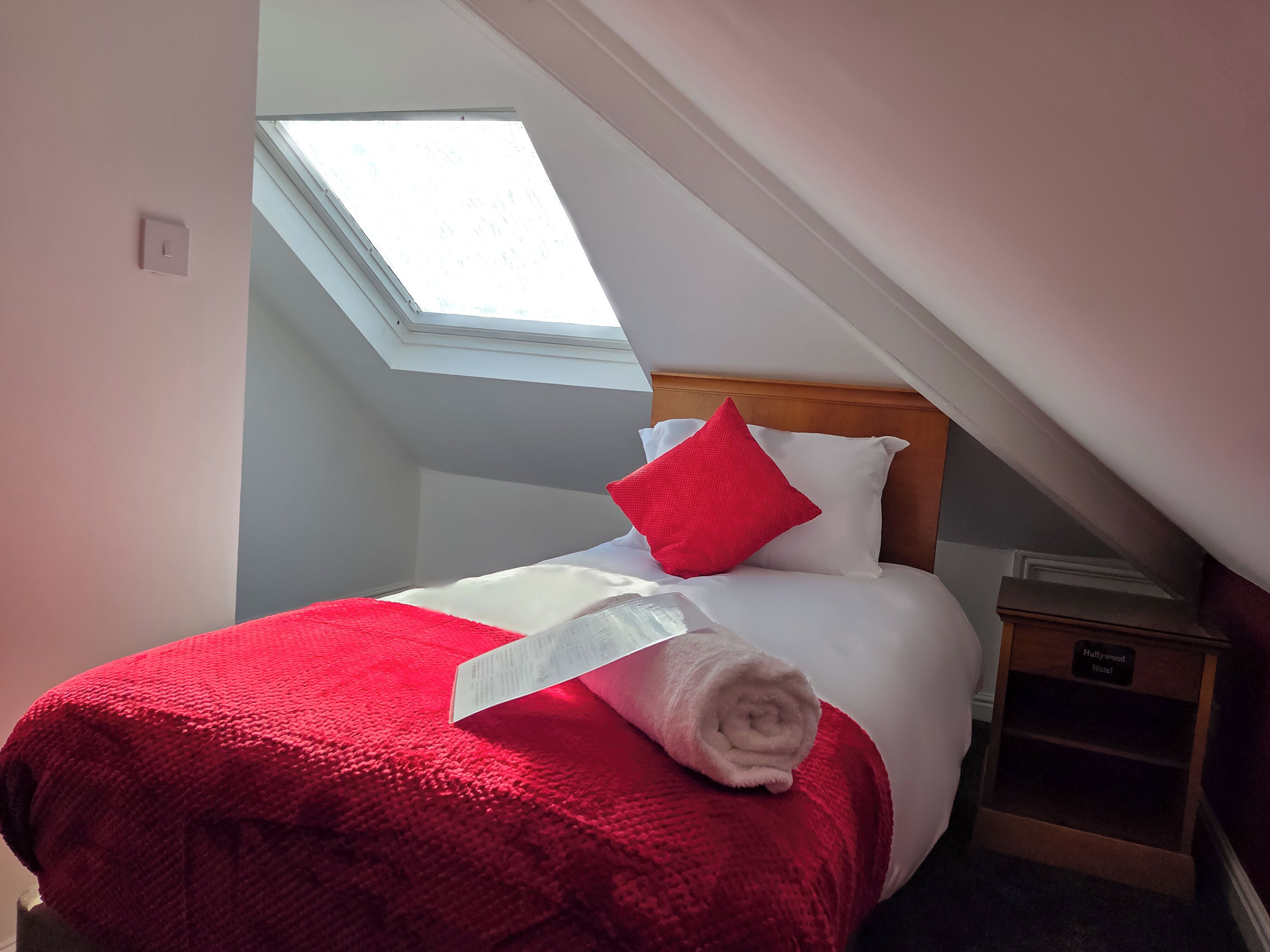 Ein ordentlich bezogenes Bett mit roter und weißer Bettwäsche, ein Oberlicht darüber und ein Nachttisch mit einem Schild, auf dem 'Hollywood Hotel' steht.