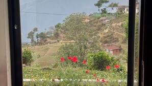 Une vue à travers une fenêtre, avec un banc rouge à l’intérieur, donnant sur une colline parsemée de fleurs rouges et sur une chaîne de montagnes lointaine.