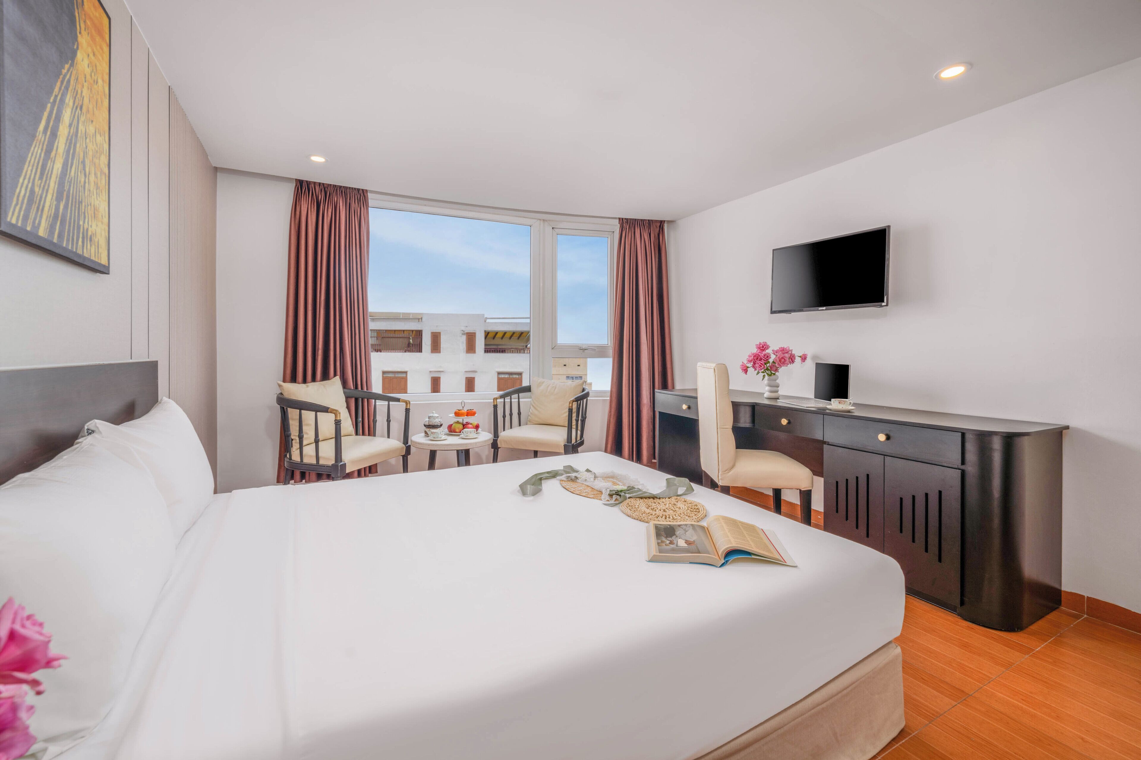 A hotel room with a large bed, a desk with a chair, a TV, and a view of buildings through the window.