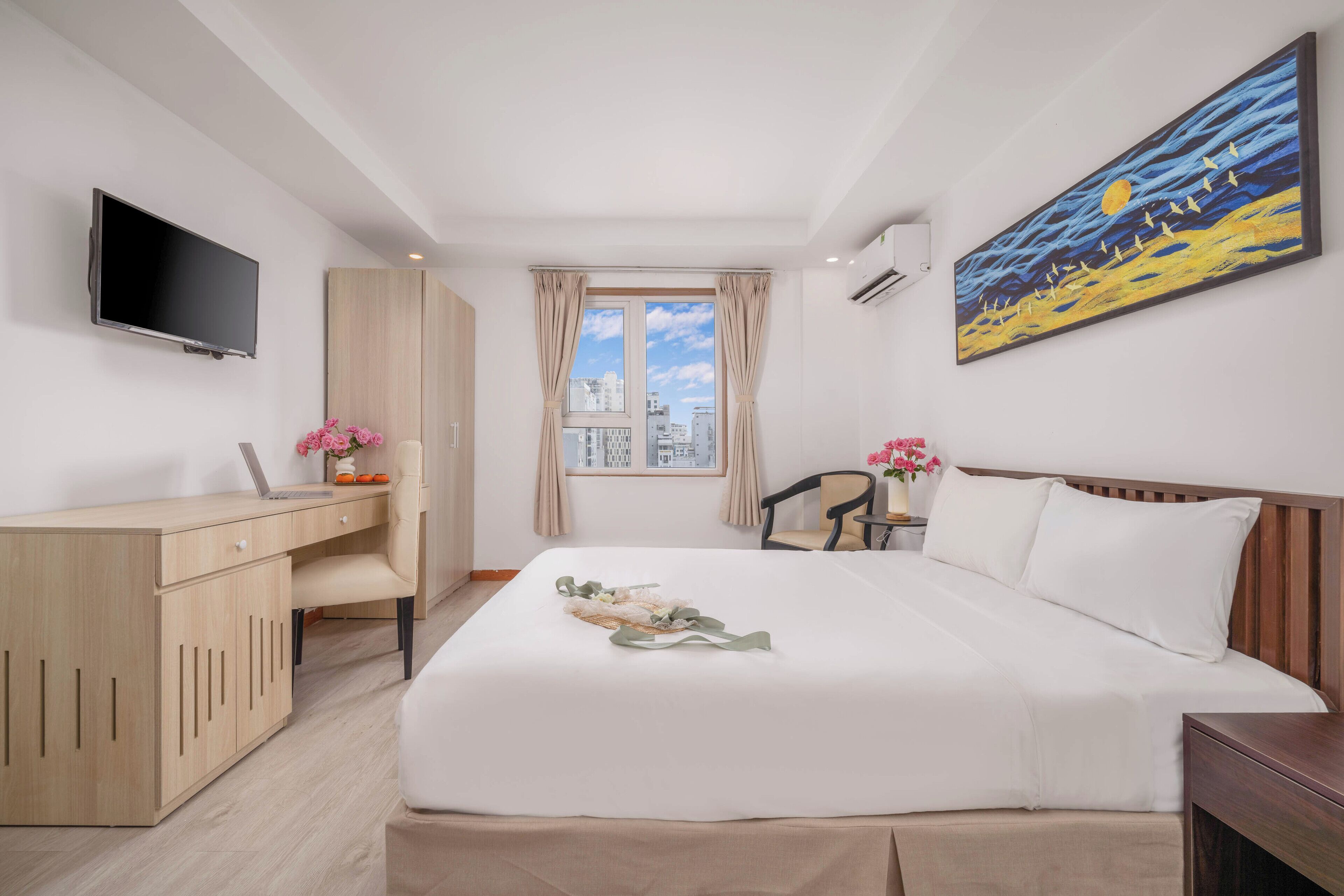 A hotel room with a bed, a desk, a TV, a window with curtains, and a painting on the wall.