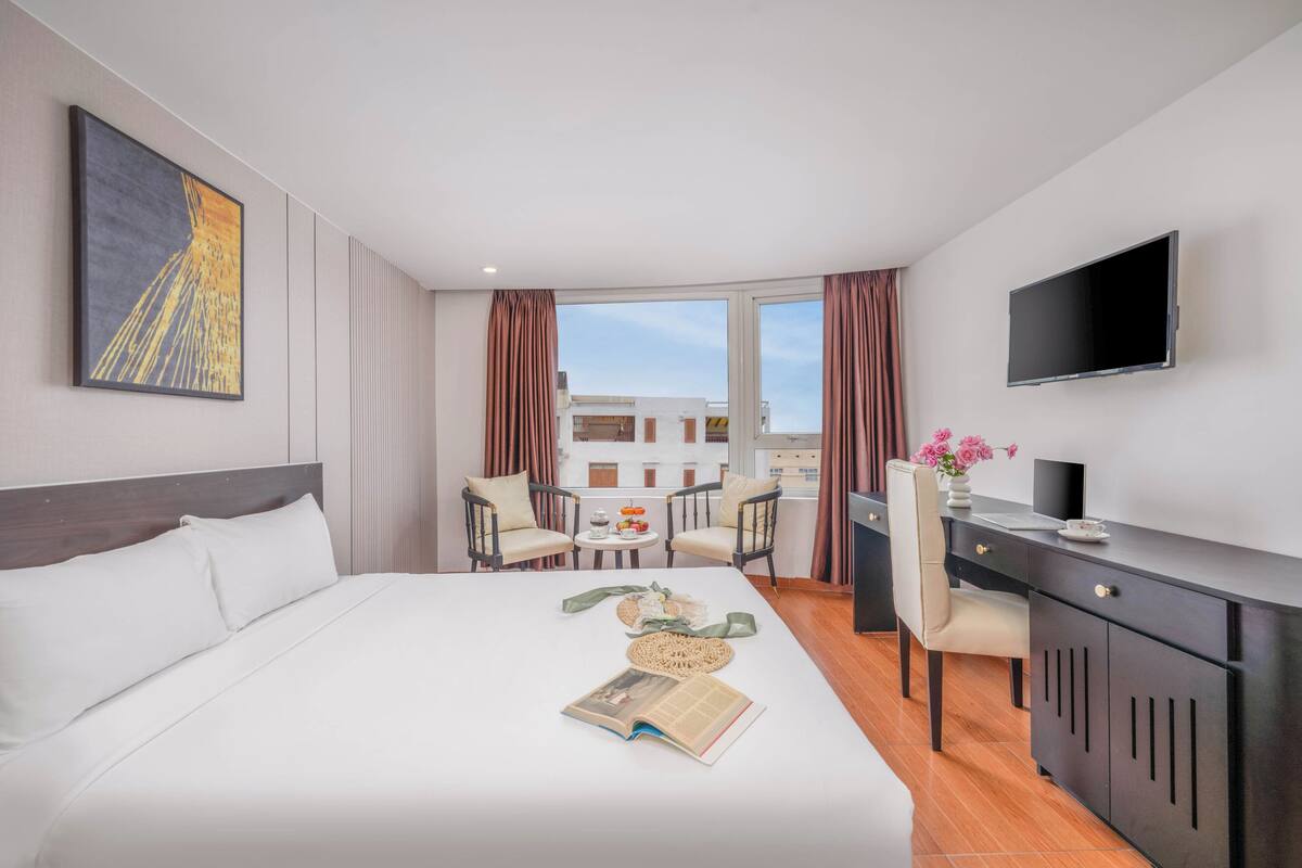 A hotel room with a bed, a desk, a chair, a TV, and a balcony with a view of buildings.