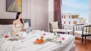 A hotel room with a bed, a laptop, a breakfast setup with fruits and flowers, and a view of buildings through the window. - Haya Hotel Da Nang (Da Nang)