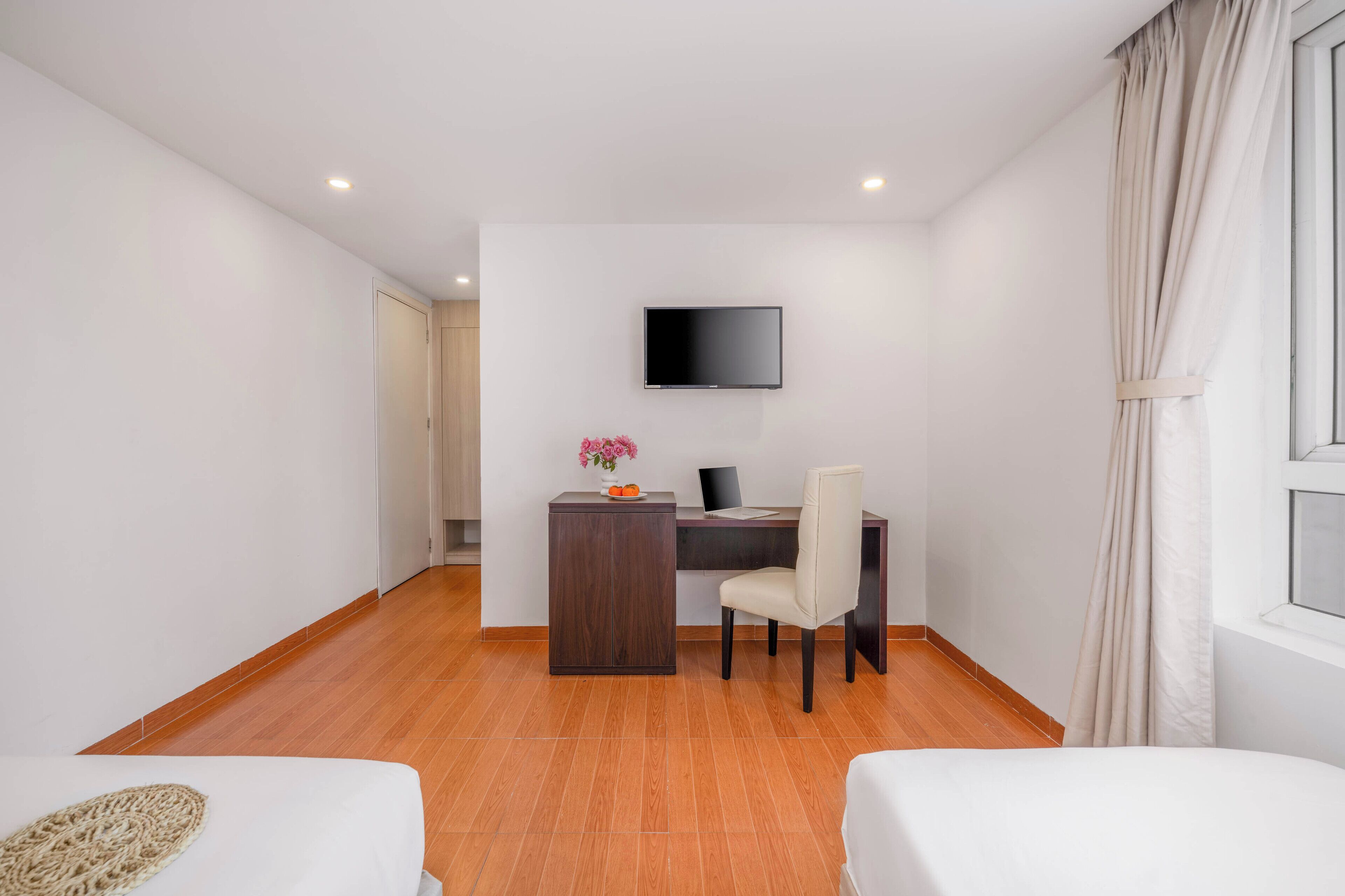 A hotel room with a wooden floor, a desk with a chair, a TV mounted on the wall, and a bed with a white cover.
