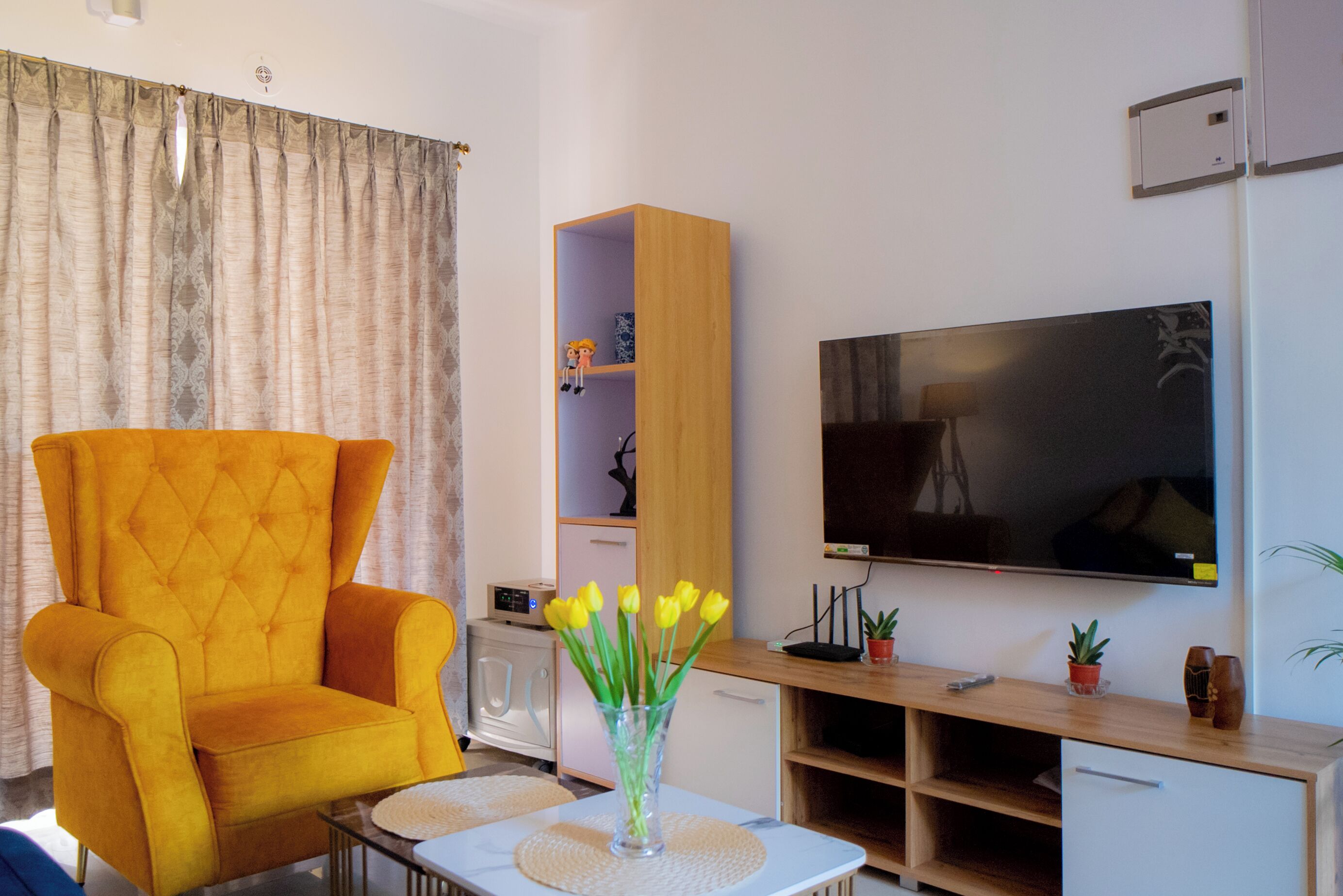 A living room with a yellow armchair, a flat-screen TV, a wooden entertainment unit, and a vase with yellow flowers.