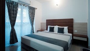 A hotel room with a bed, a nightstand, a wooden headboard, and a window with curtains. - Ocean Whisper Goa (Goa)