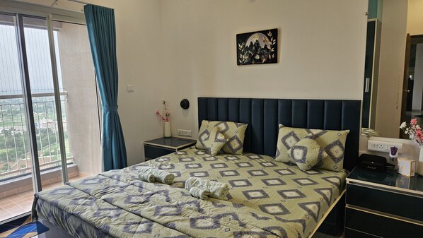 Una habitación de hotel con cama, mesita de noche y un reloj en la pared. Se ve una ventana con cortina y vistas al exterior.
