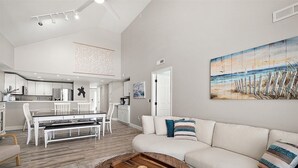 A modern living room with a white sofa, a dining area with white chairs, and a kitchen with white cabinets. - Aventurra 403 — Modern Beach Block Penthouse Oasis with Rooftop Pool, Bay Views & Walkable Fun — Sleeps 9 (Ocean City)