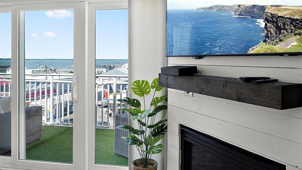 A room with a large TV mounted on the wall, a fireplace, and a view of the ocean through large windows. - Aventurra 403 — Modern Beach Block Penthouse Oasis with Rooftop Pool, Bay Views & Walkable Fun — Sleeps 9 (Ocean City)