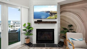 A modern living room with a fireplace, a large TV mounted above it, and a view of the ocean through a large window. - Aventurra 403 — Modern Beach Block Penthouse Oasis with Rooftop Pool, Bay Views & Walkable Fun — Sleeps 9 (Ocean City)
