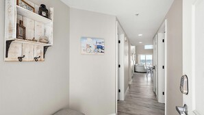 A hallway with a bench, a wall-mounted shelf with decorative items, and a painting of a bicycle. - Aventurra 403 — Modern Beach Block Penthouse Oasis with Rooftop Pool, Bay Views & Walkable Fun — Sleeps 9 (Ocean City)